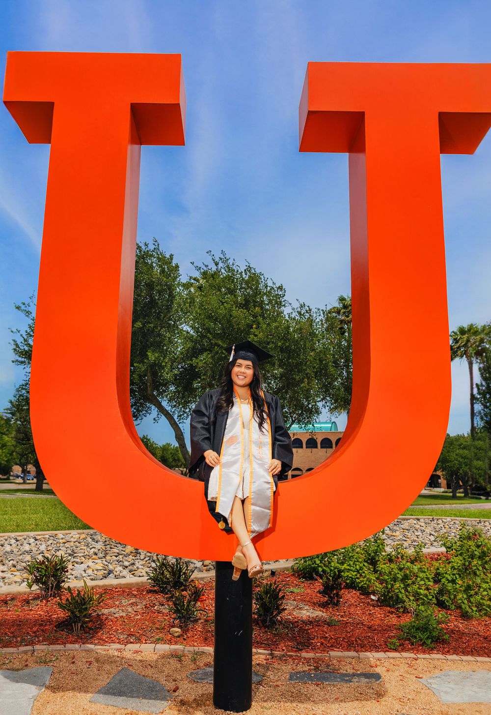 Graduate in academic regalia poses at large red U-shaped university sign outdoors.