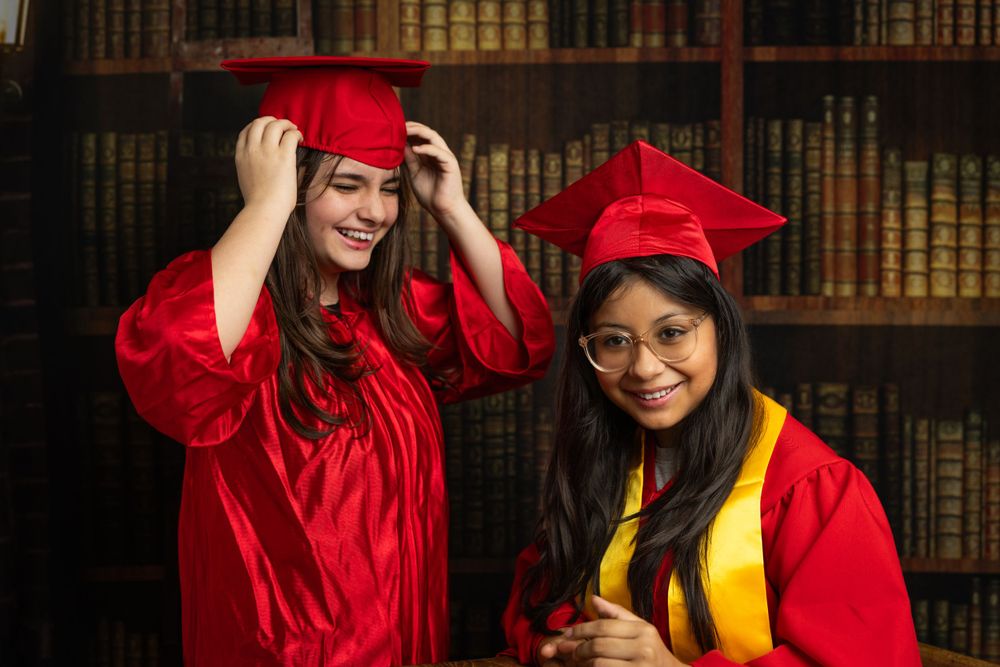 Two graduates in red caps and gowns share a joyful moment in front of library bookshelves.