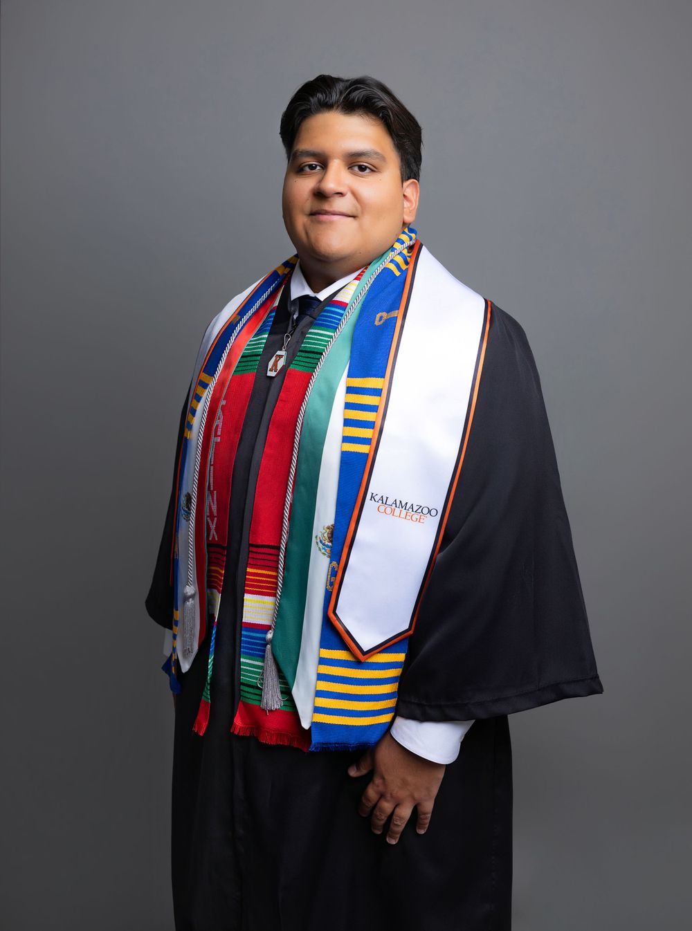 Graduate wearing traditional academic regalia with colorful stoles and sashes against a gray background.