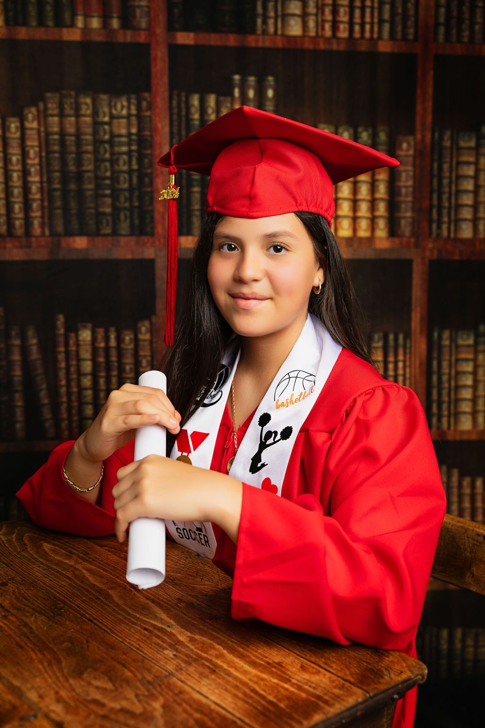 Graduate holding diploma while posing in red cap and gown against library background.