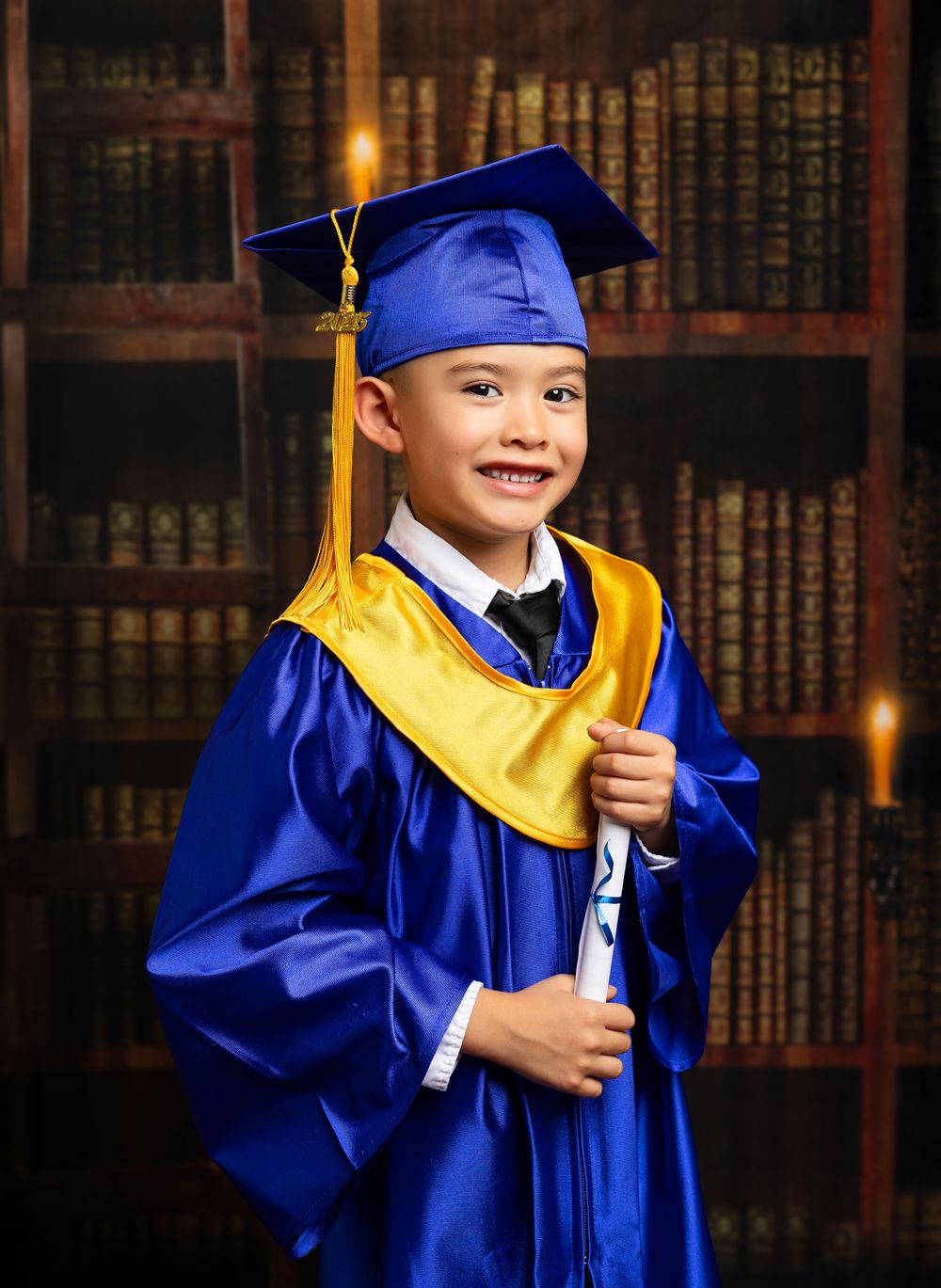 Young graduate in blue and yellow cap and gown holding diploma smiles in front of library bookshelves.