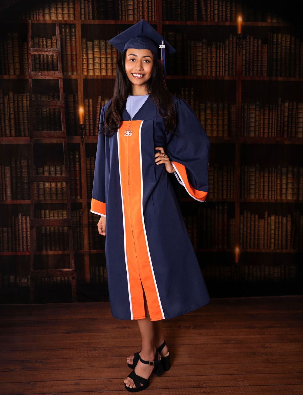 Young woman in navy blue and orange graduation gown and cap smiling in front of a library bookshelf backdrop.