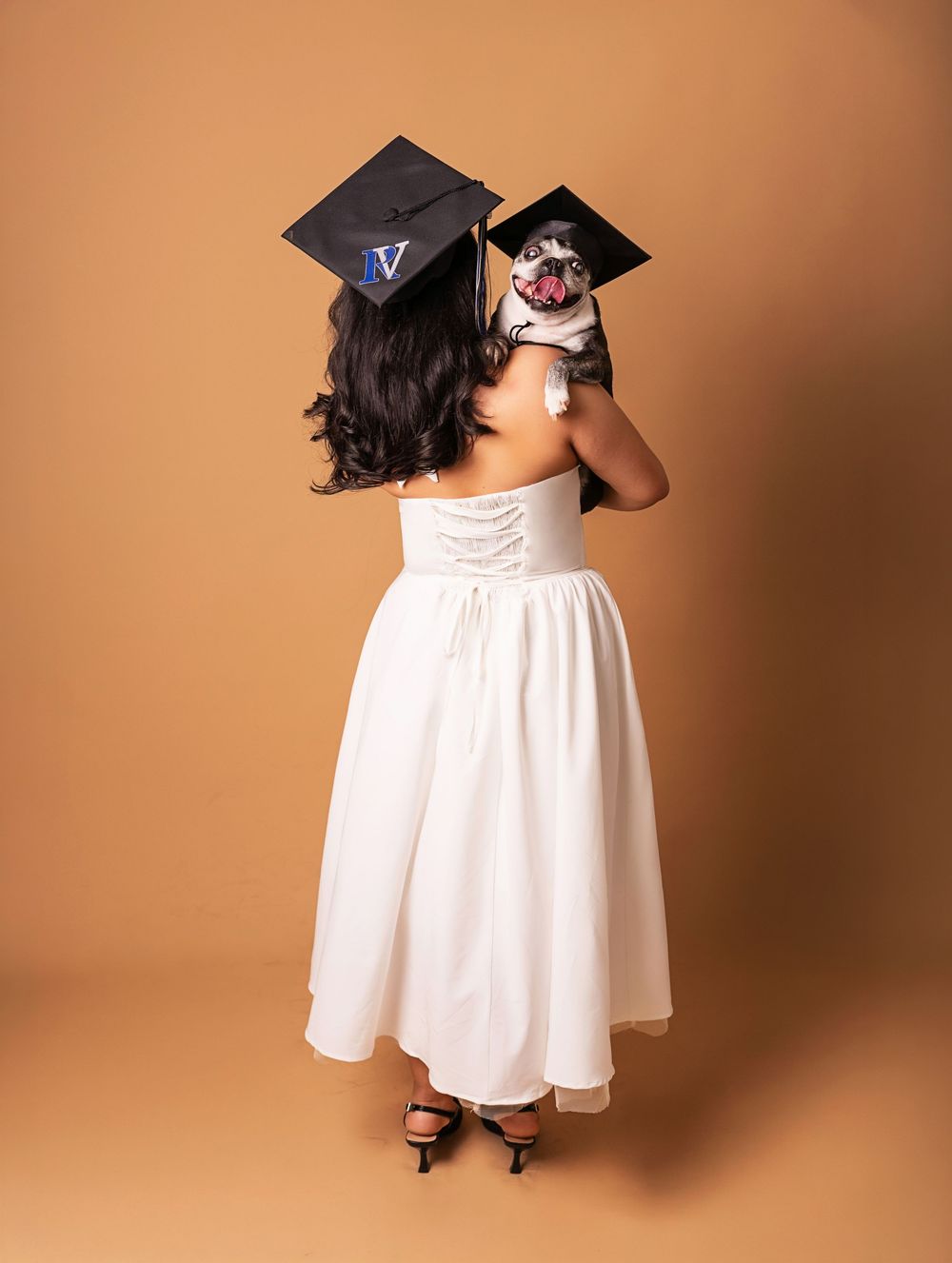 Woman in white dress and graduation cap holds a dog wearing a matching cap, posing for a grad photo.