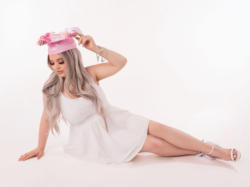 Woman in white dress and pink floral hat poses gracefully on white studio floor with silver heels.