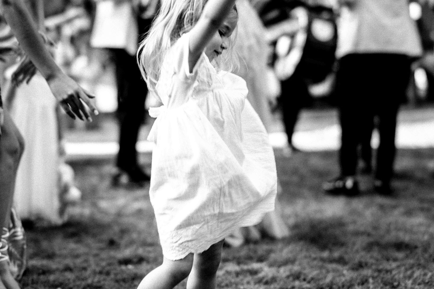 Black and white photo of a child twirling in a white dress while dancing outdoors among a crowd.