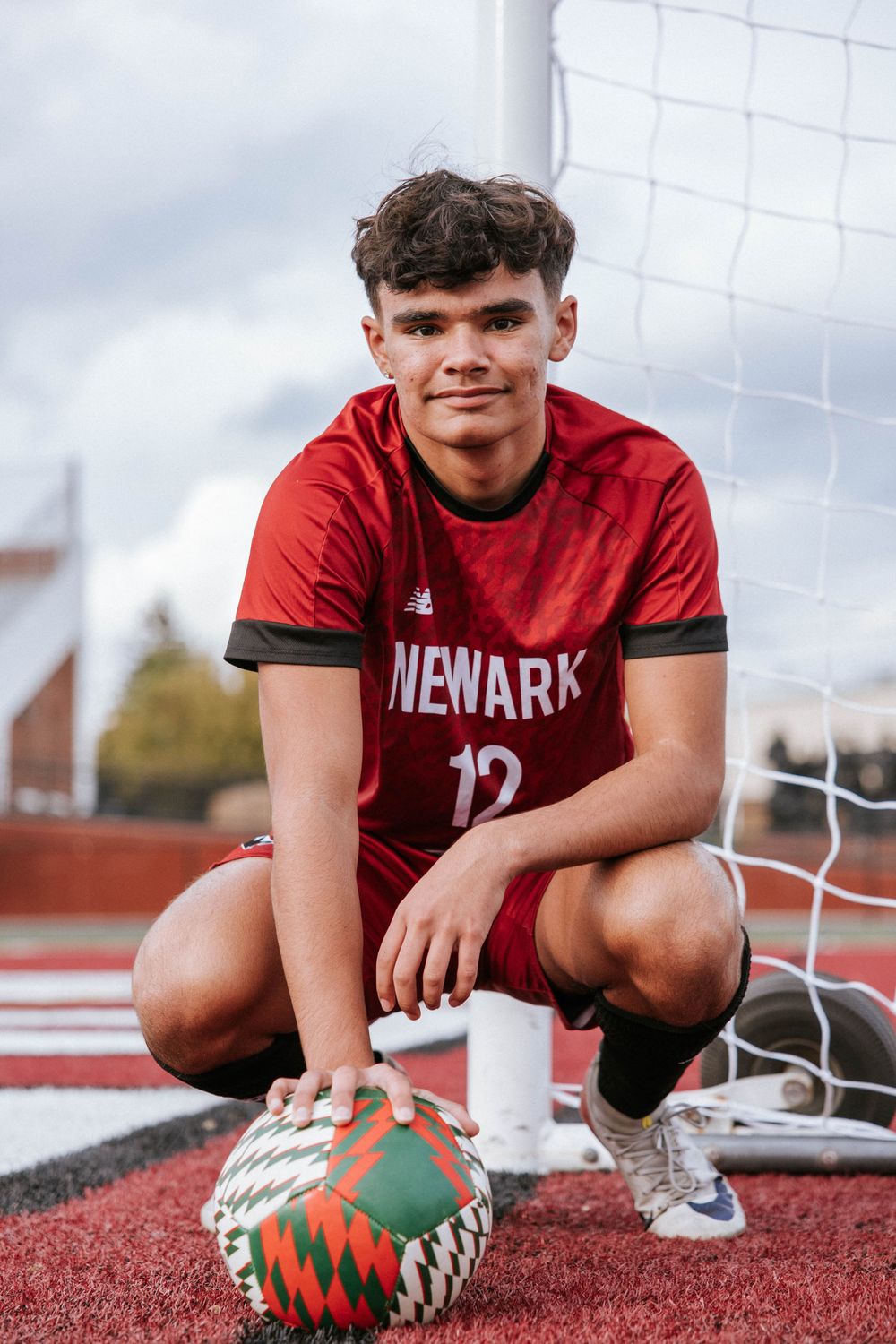 Soccer player in red Newaha jersey poses with soccer ball near goal net on red turf field.