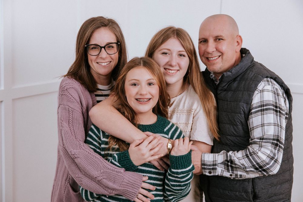 A family wearing coordinated green and plaid outfits embraces while posing for holiday photos in their home.