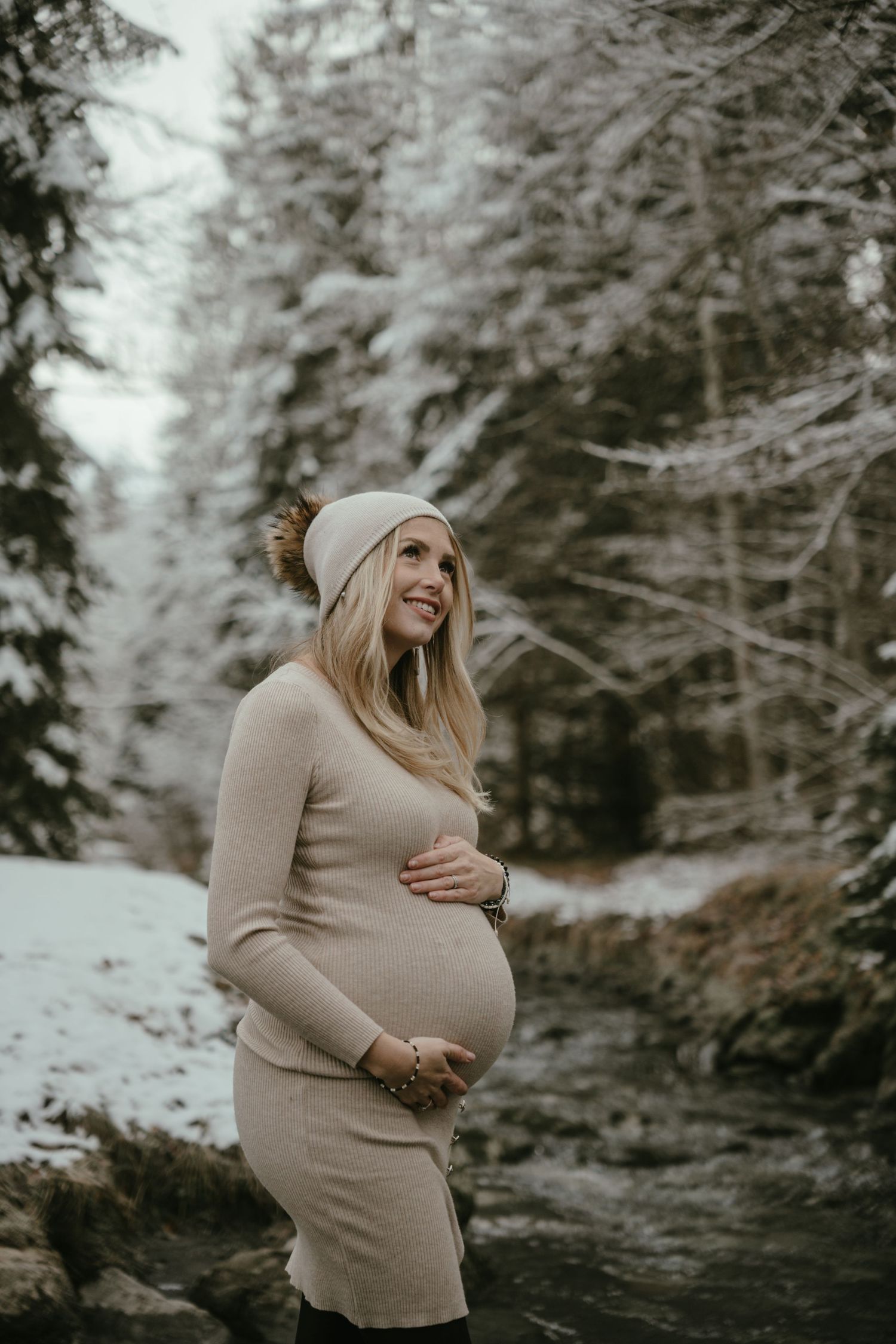 Expectant mother in beige sweater dress poses in snowy winter forest setting.