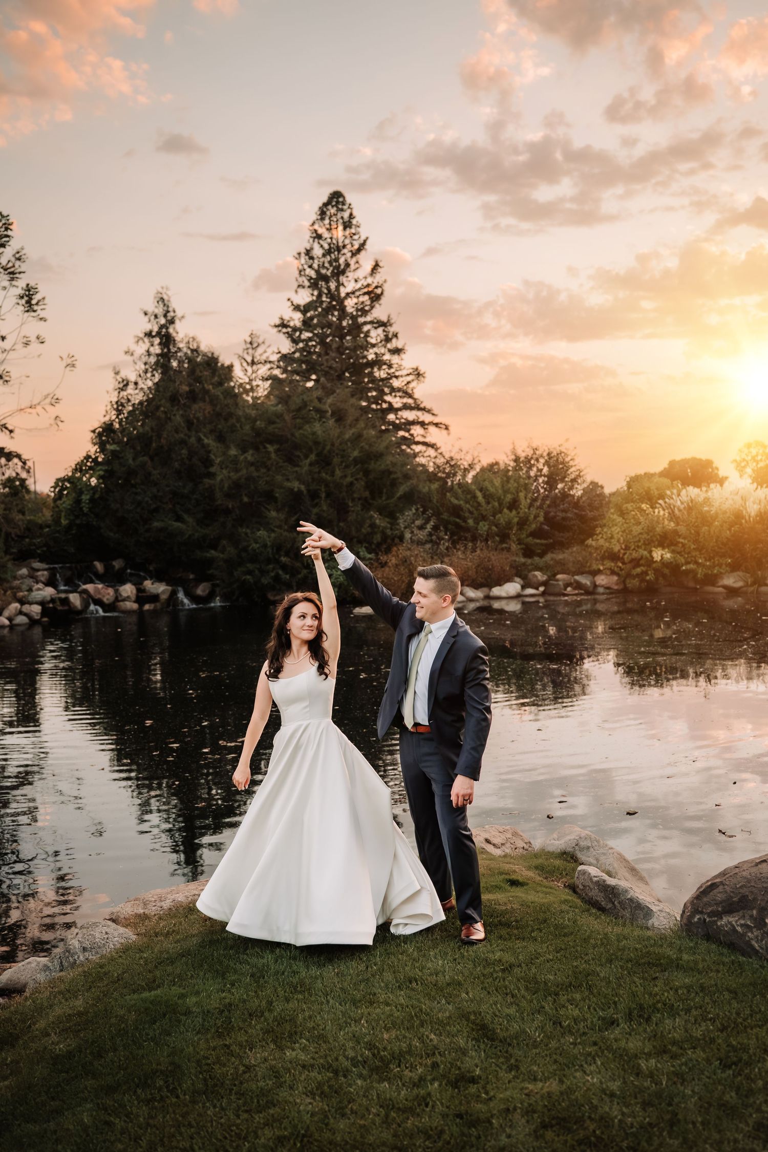 Bride and groom dancing by a serene pond at sunset, surrounded by lush trees and golden light.