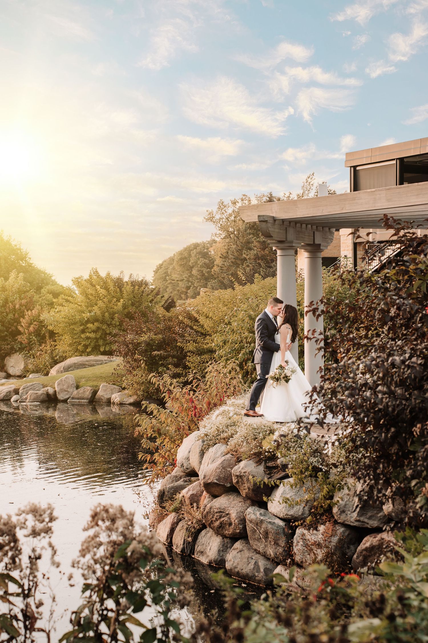 A couple stands in an elegant gazebo overlooking a serene pond with rocks and foliage during golden hour sunset.