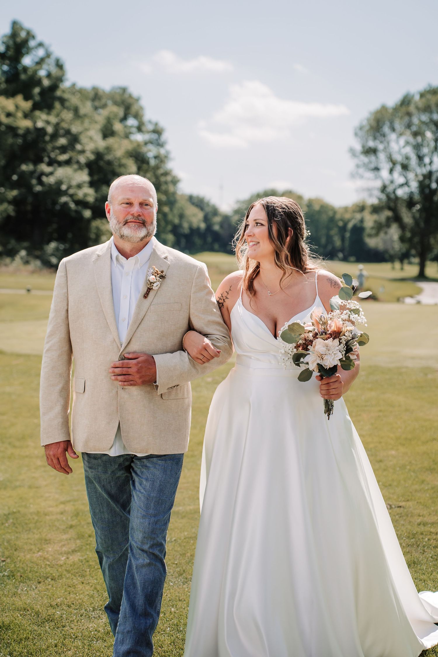 Bride in white gown holding bouquet walks arm-in-arm with older man in tan blazer and jeans on green lawn.