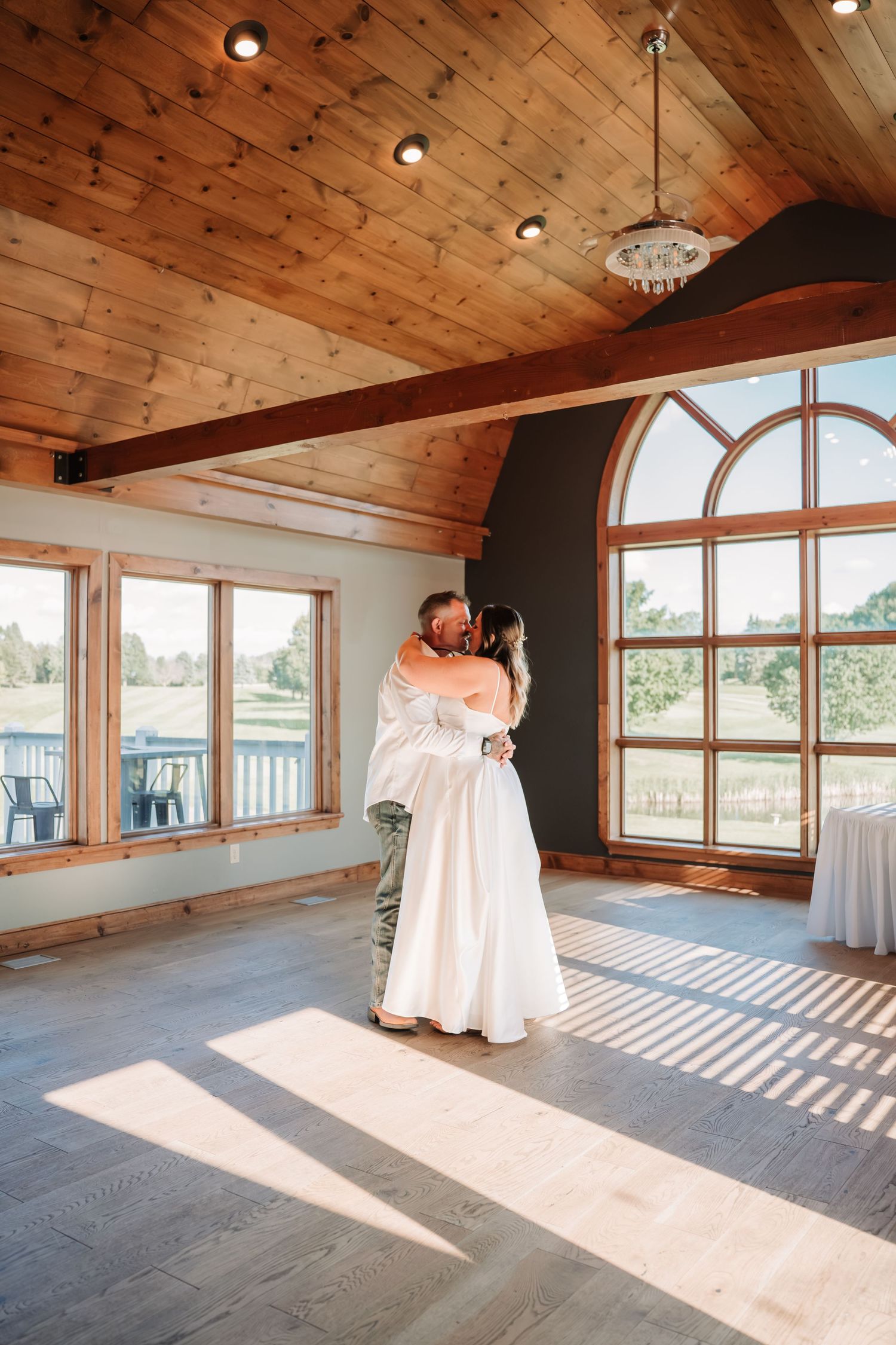 Bride and groom share their first dance in a rustic wooden venue with arched windows and warm natural light.