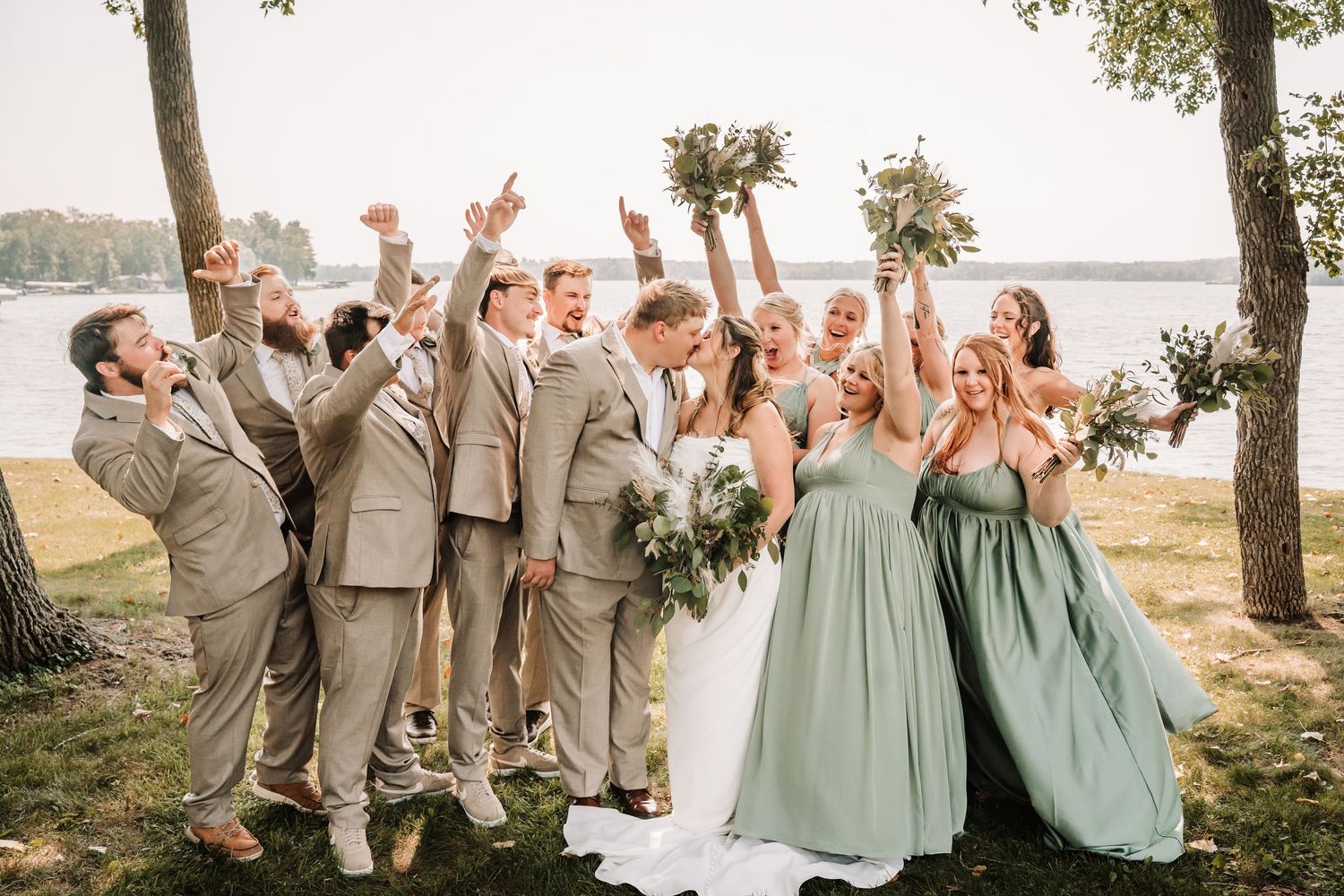 Wedding party in sage green dresses and tan suits celebrate joyfully outdoors near a waterfront setting.