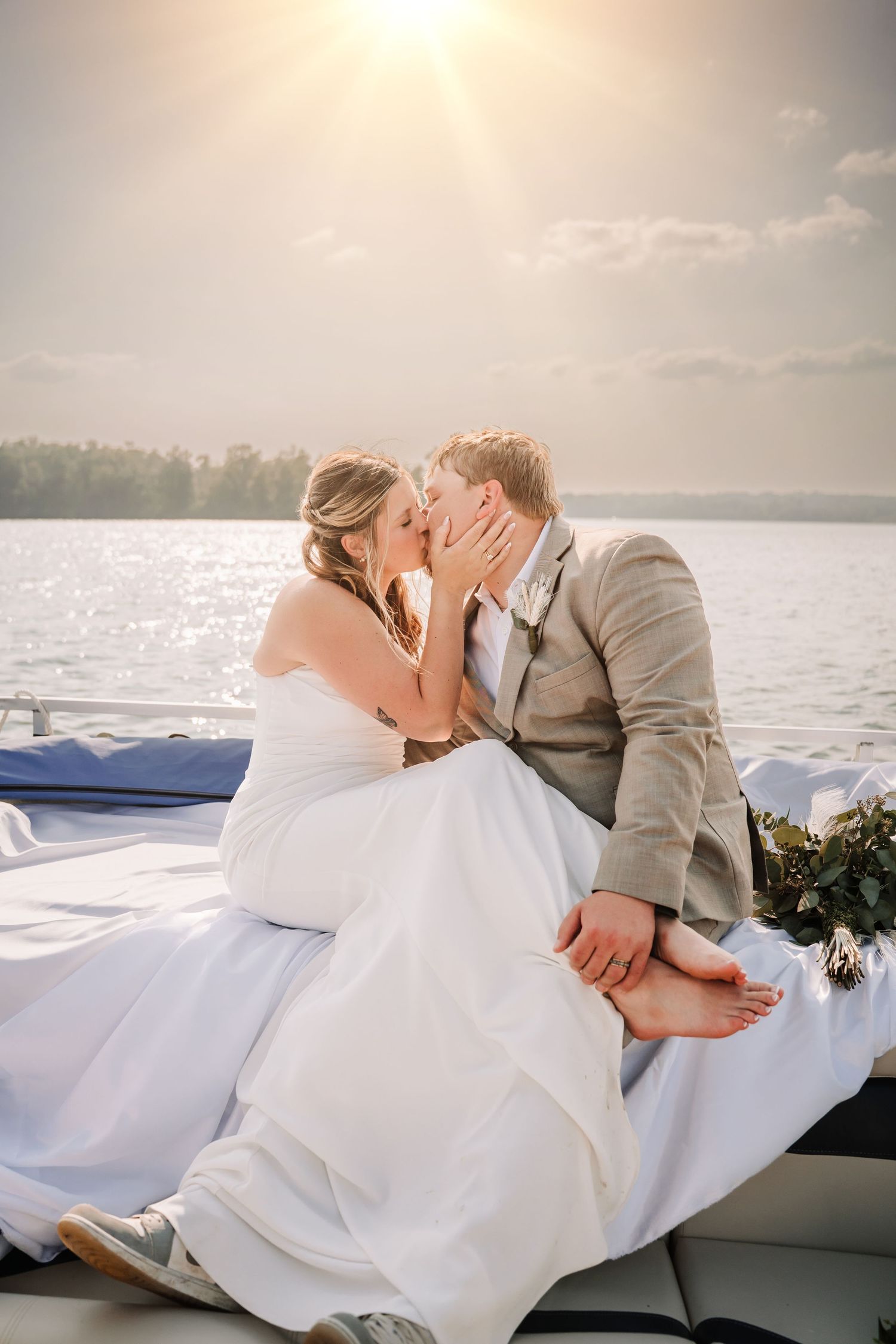 A romantic wedding couple shares a kiss on a boat at sunset with a lake and sunbeams in the background.