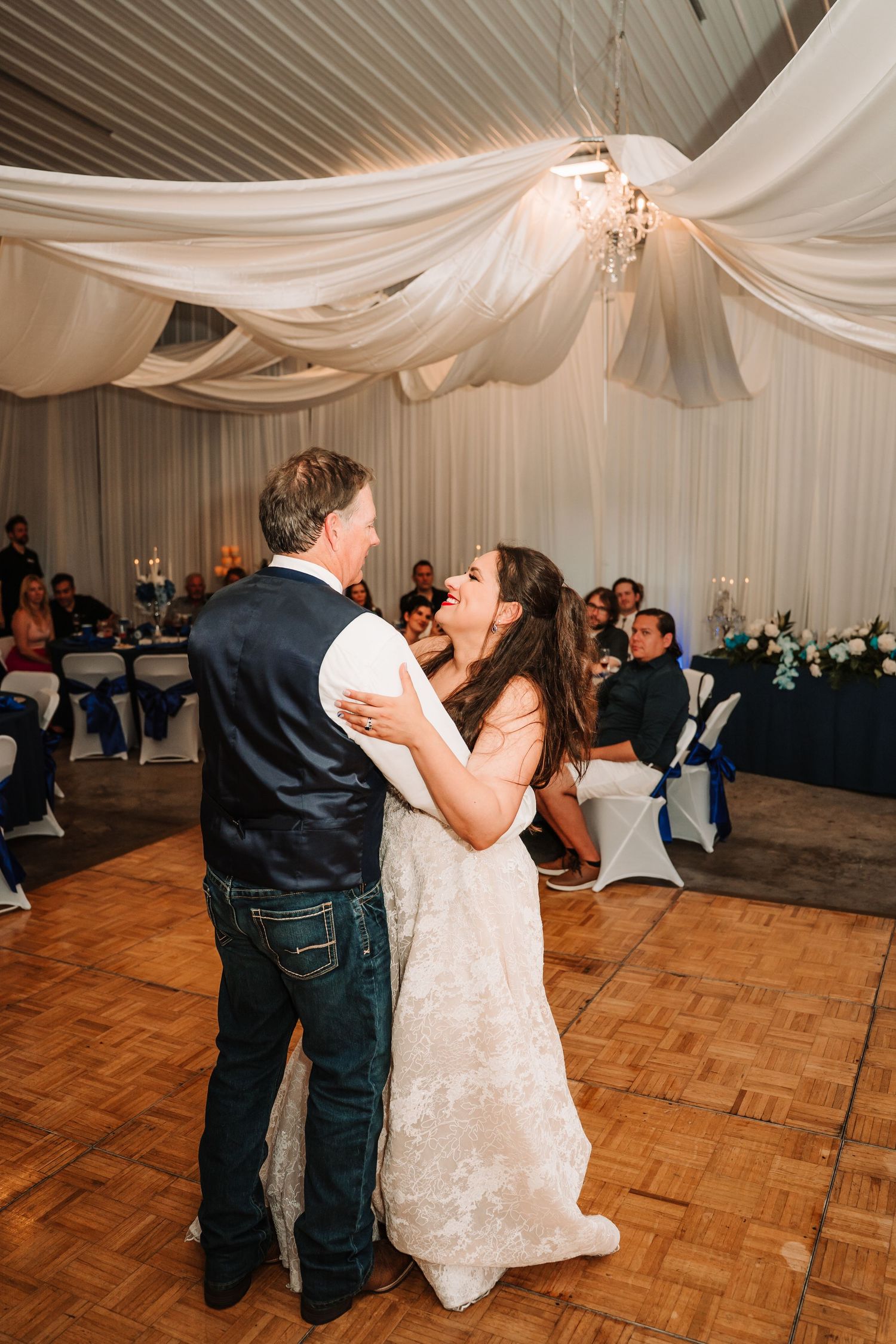 A couple shares their first dance as newlyweds on a wooden dance floor under draped white fabric at their wedding reception.