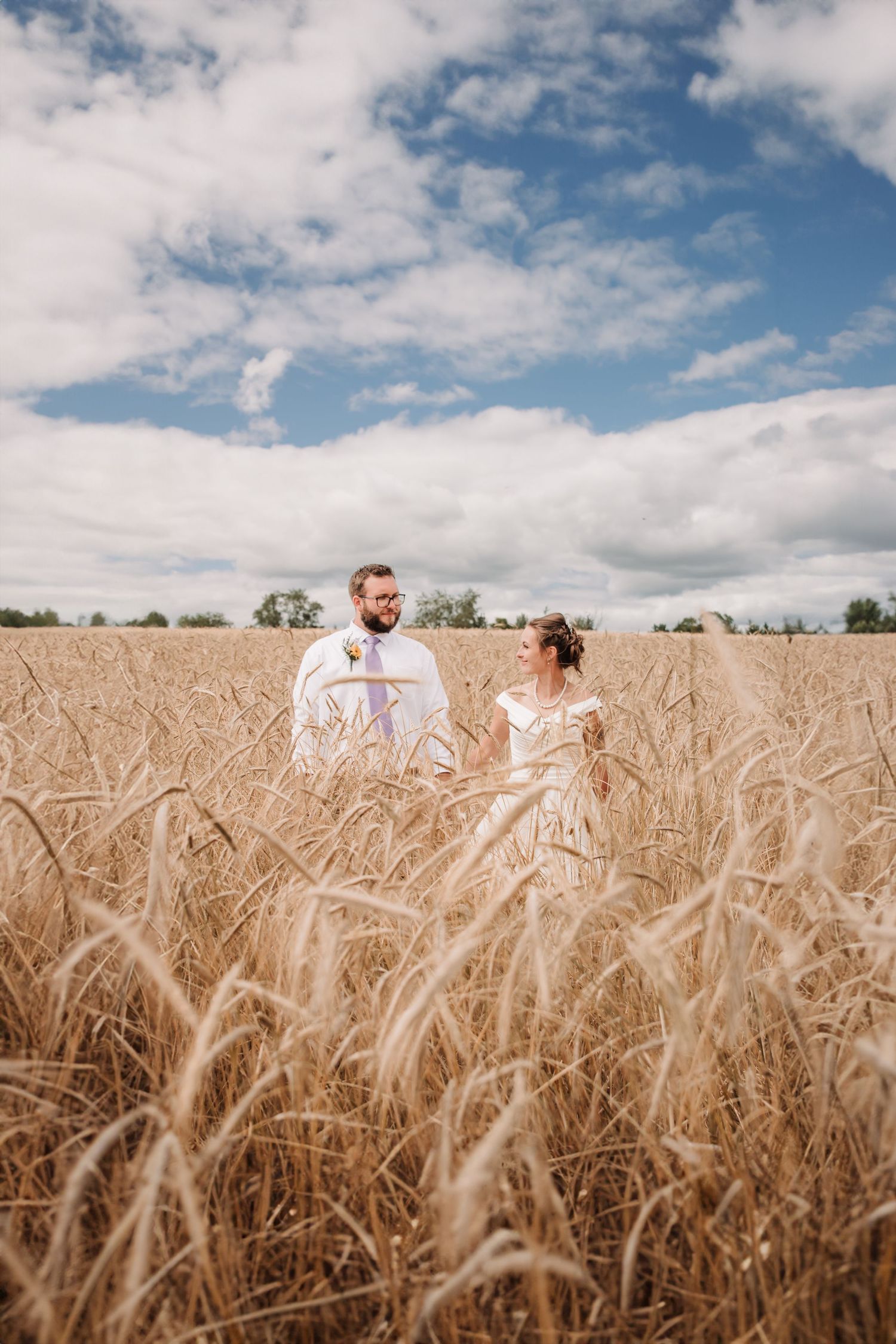 Romantic couple stands in golden wheat field under cloudy sky during countryside photoshoot.