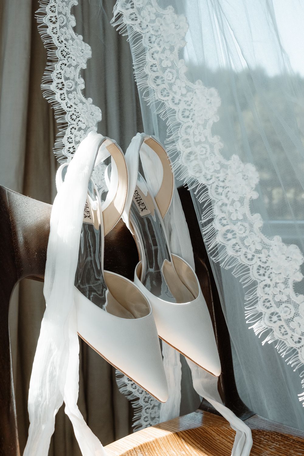 Elegant white slingback wedding heels sitting on a wooden surface near delicate lace curtains.