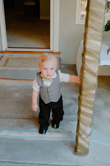 Small child in formal gray vest and black pants crawls on gray concrete floor near decorative column.