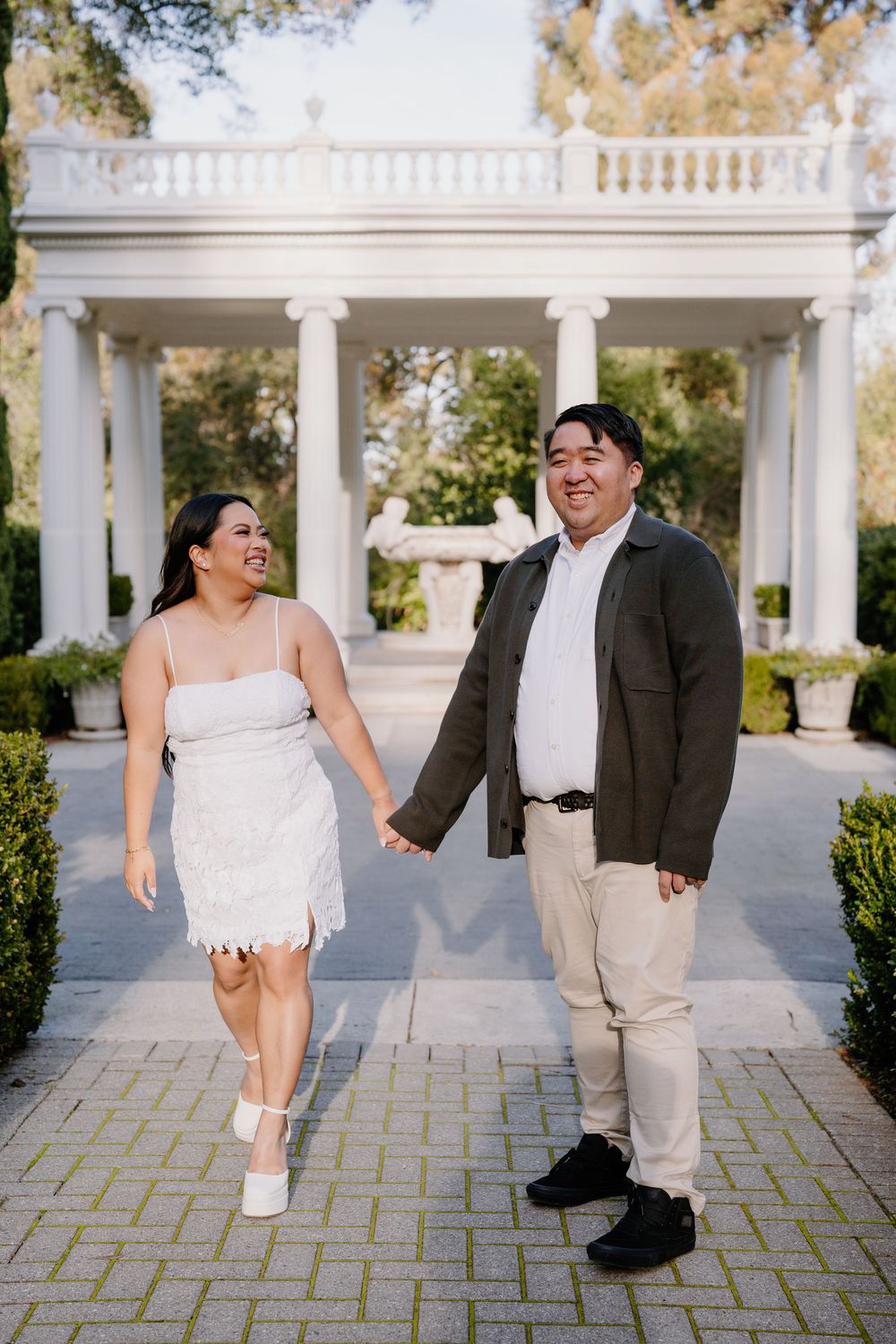 A couple in wedding attire shares playful moments together under white columns on a stone patio.