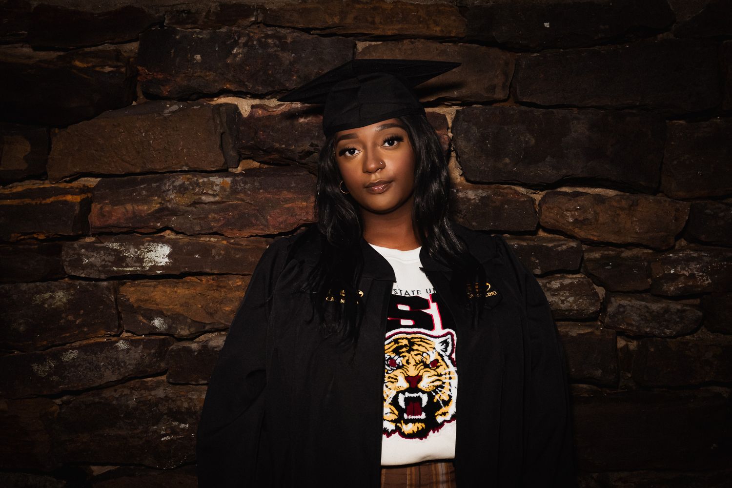 A graduate wearing a black jacket and graduation cap poses against a rustic brick wall background.
