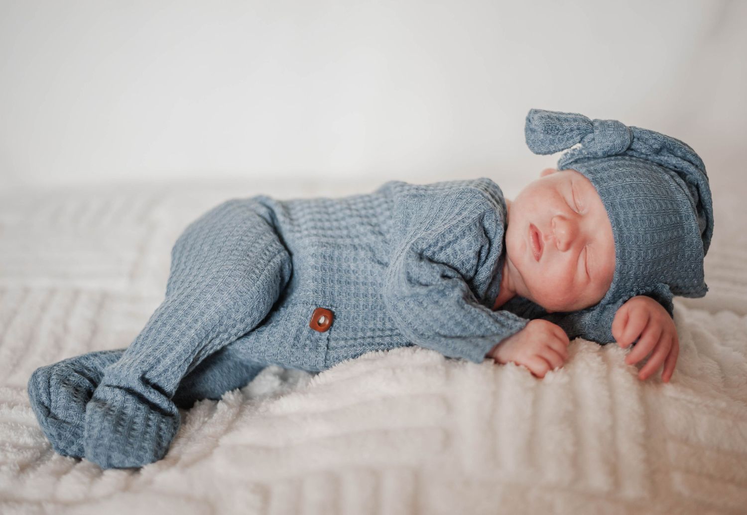 A sleeping baby wearing a soft blue knitted romper outfit with buttons lies on white bedding.