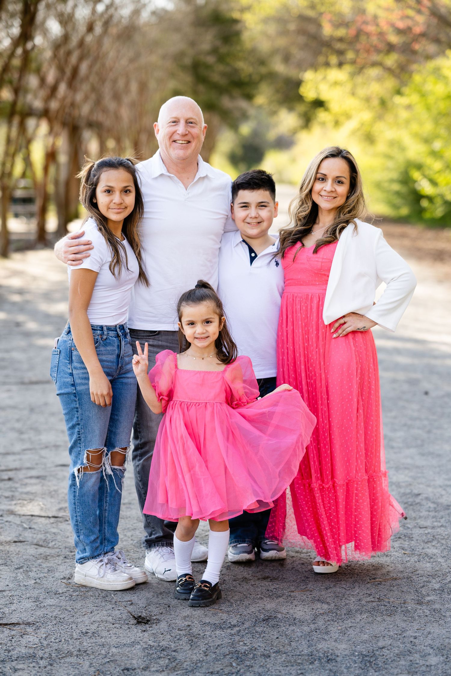 Family of five wearing coordinating pink and white outfits pose together on a tree-lined path.
