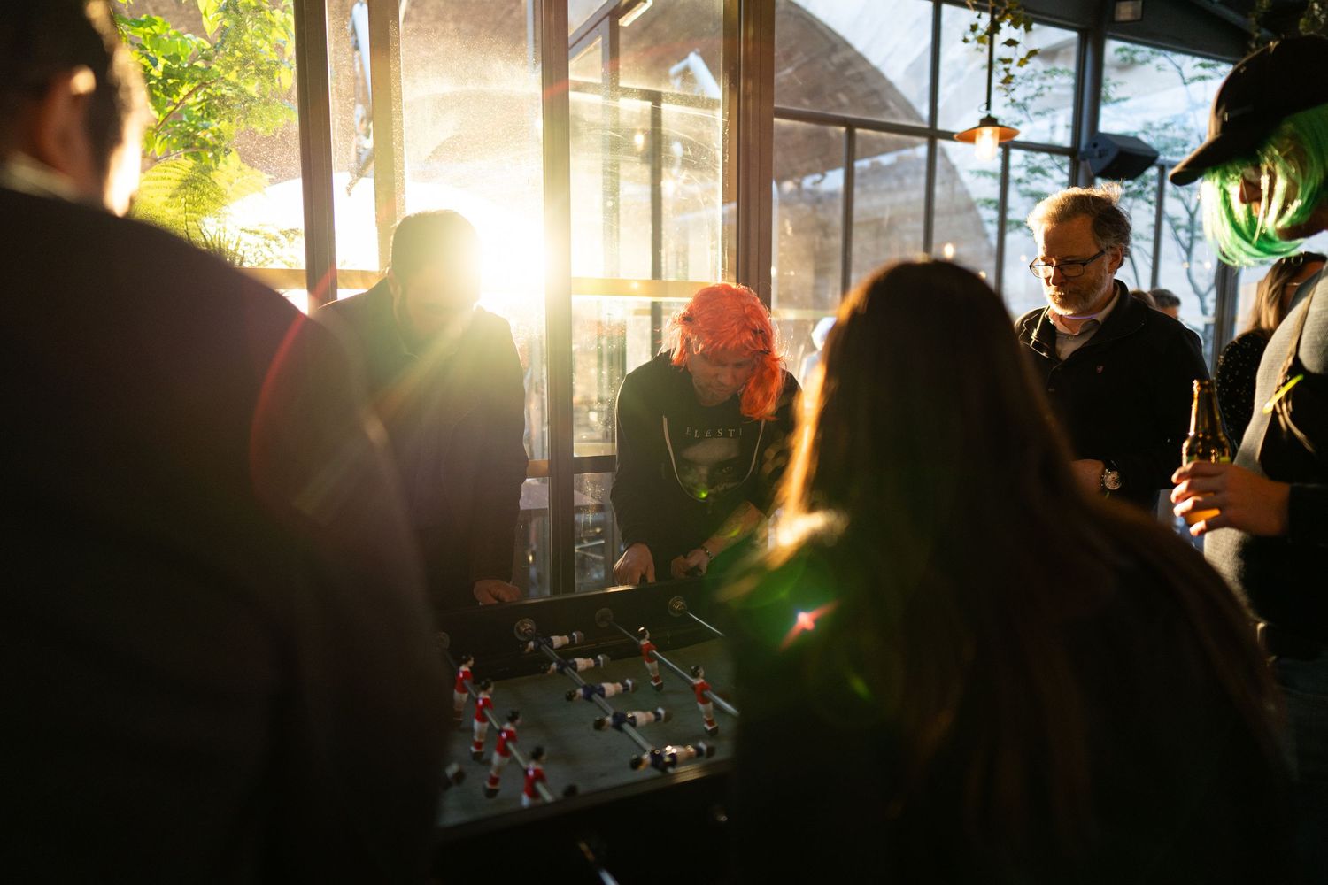 A crowd of people gathered around a table in a sunlit room with warm lens flare streaming through the windows.