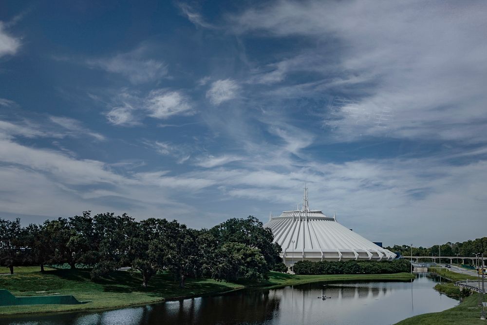 Space Mountain dome building reflected in a lake at Disney World with blue sky and wispy clouds.