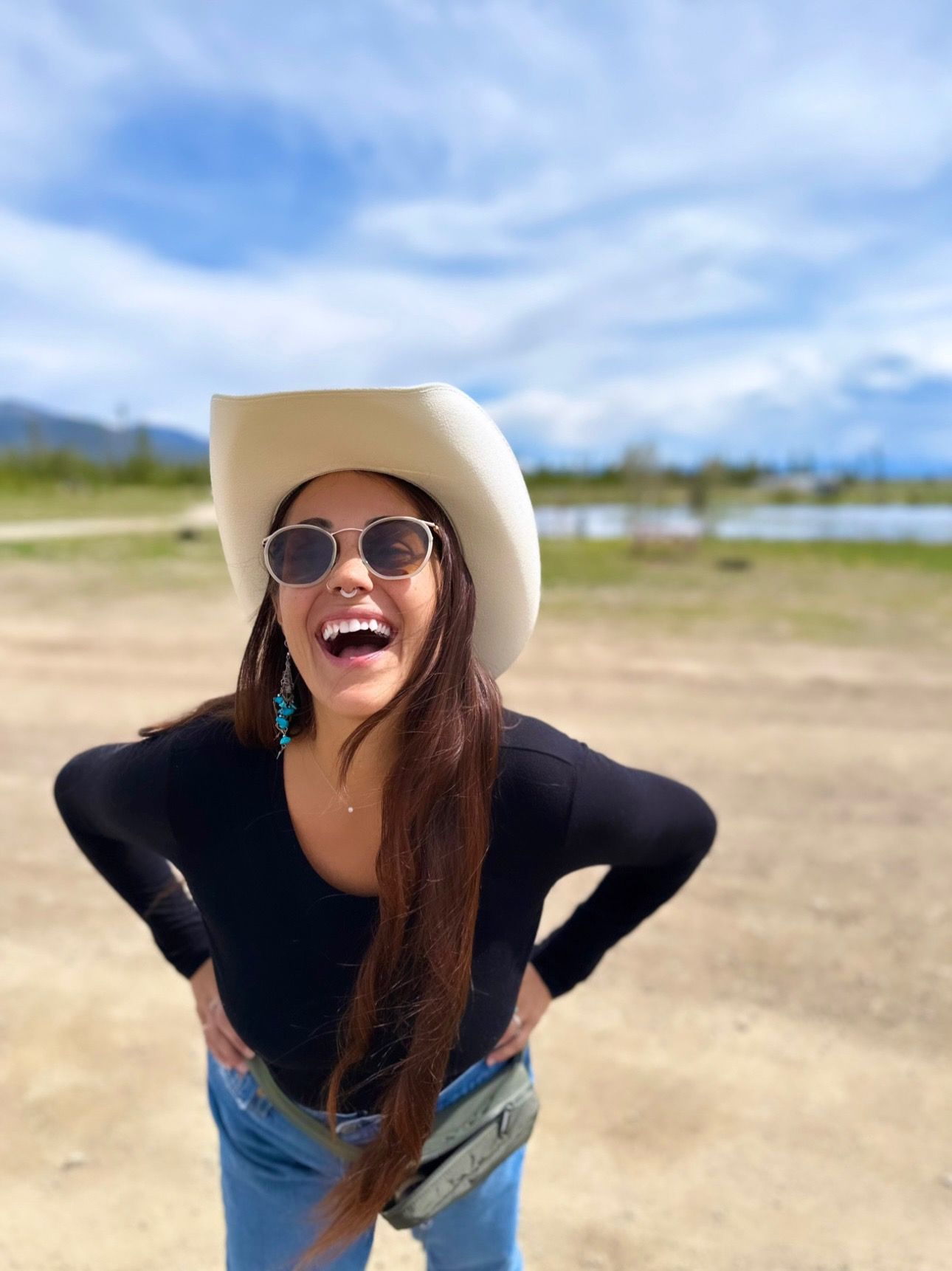Happy woman wearing a cowboy hat, sunglasses, and turquoise earrings laughing outdoors under a blue sky.