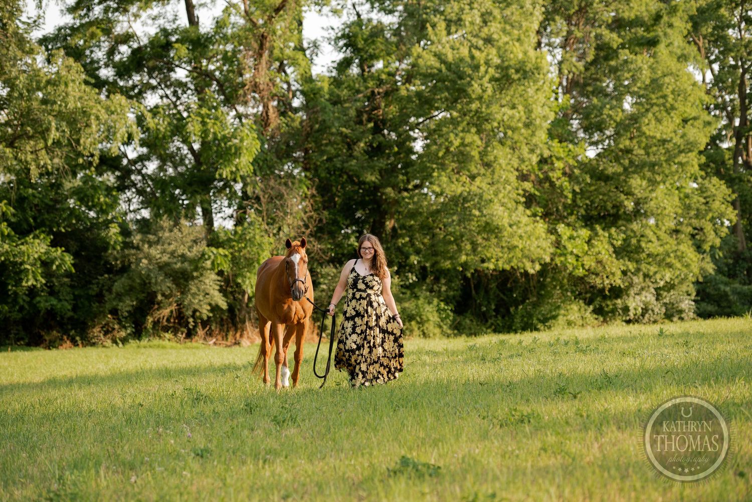 A chestnut horse and someone in a floral dress walk together in a sunlit field.