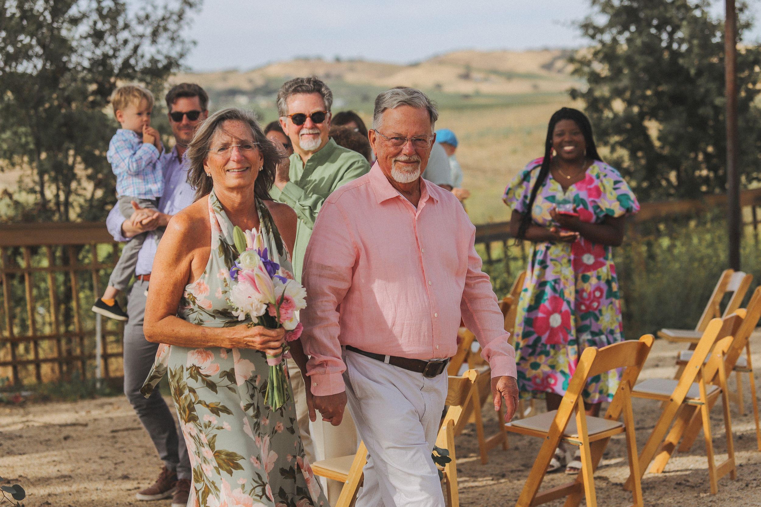 A celebratory outdoor gathering with people in formal attire walking past folding chairs.