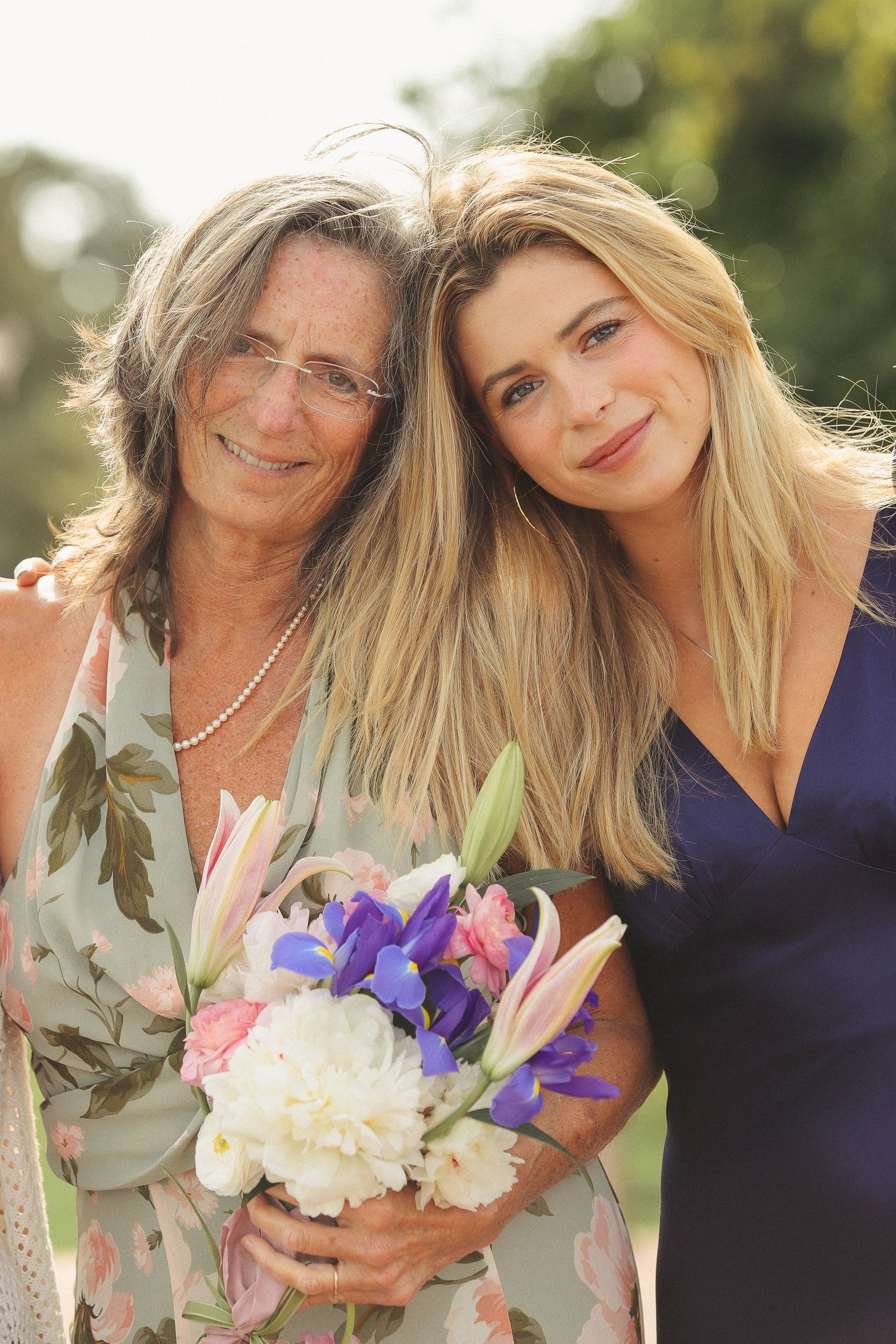 Two people wearing floral and navy dresses posing with a bouquet of purple and white flowers outdoors.