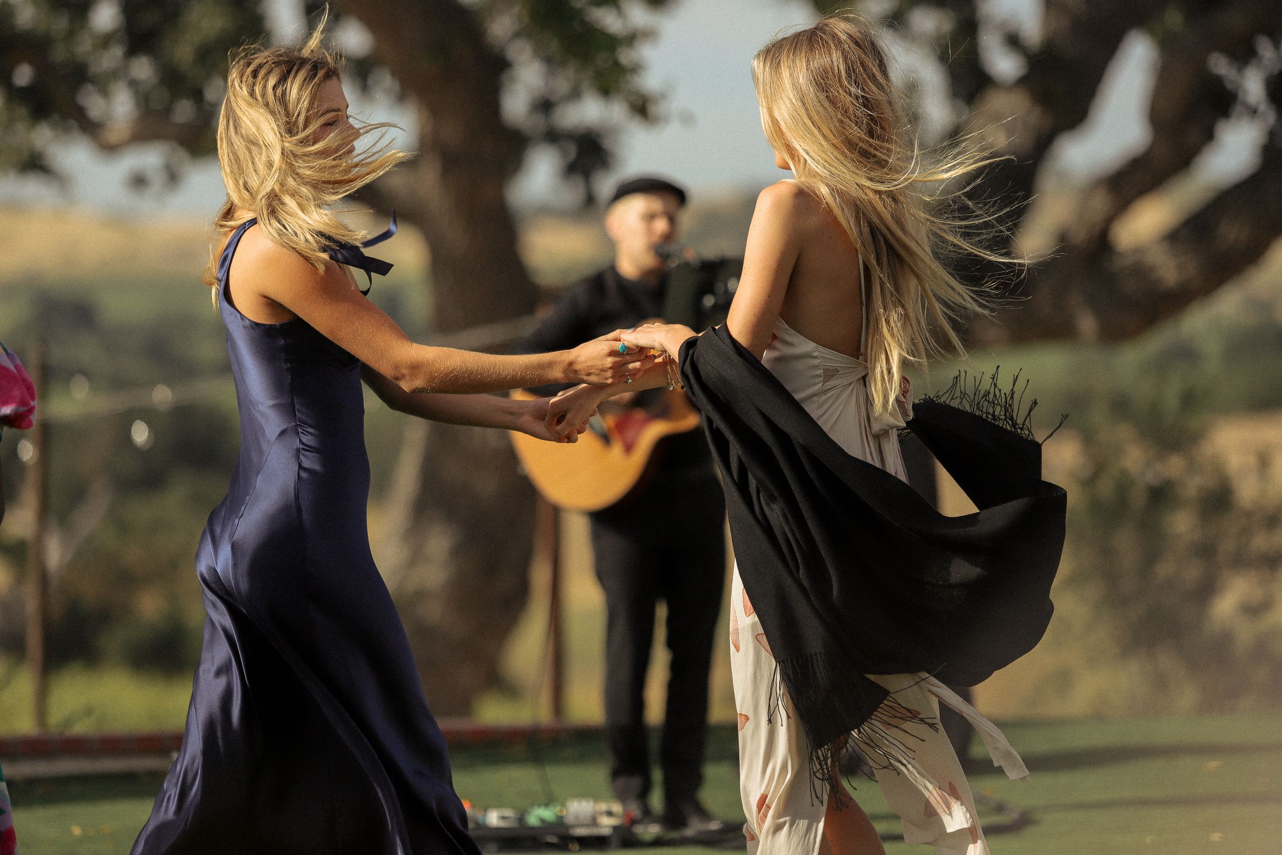 Two people in flowing dresses dancing together at an outdoor event with a musician in the background.