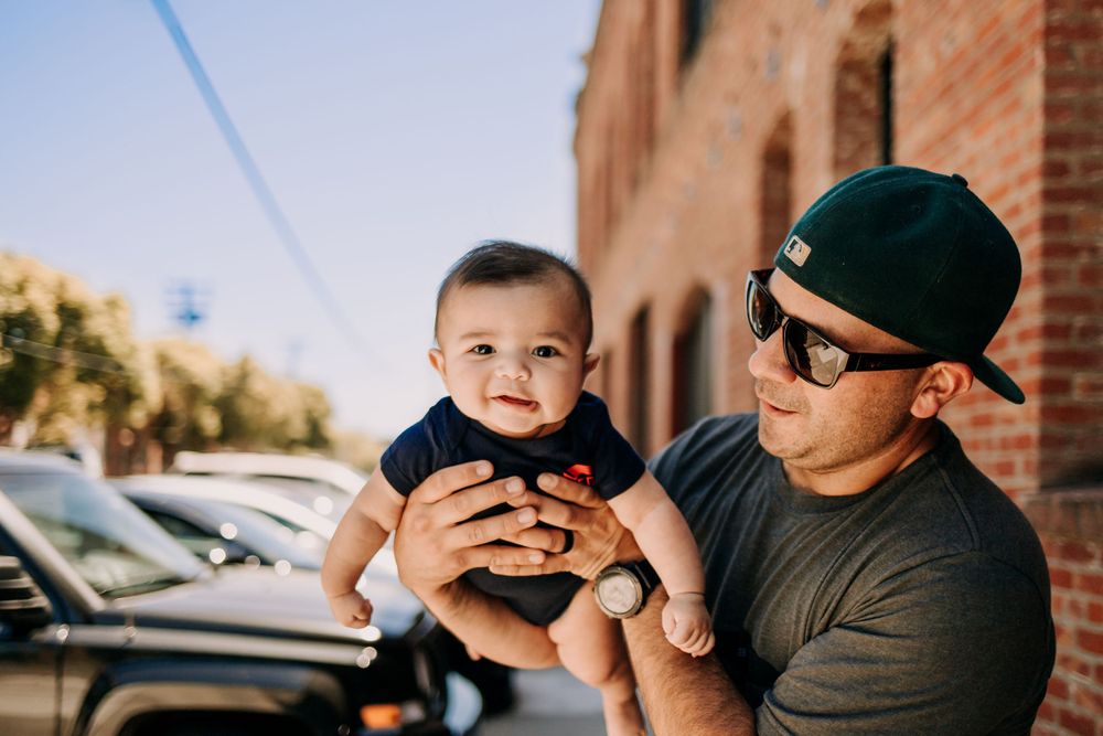 Adult holding baby outdoors near brick building on sunny day.