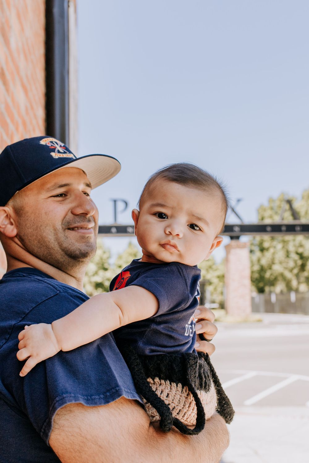 Baby being held by adult wearing baseball cap outdoors near brick building.