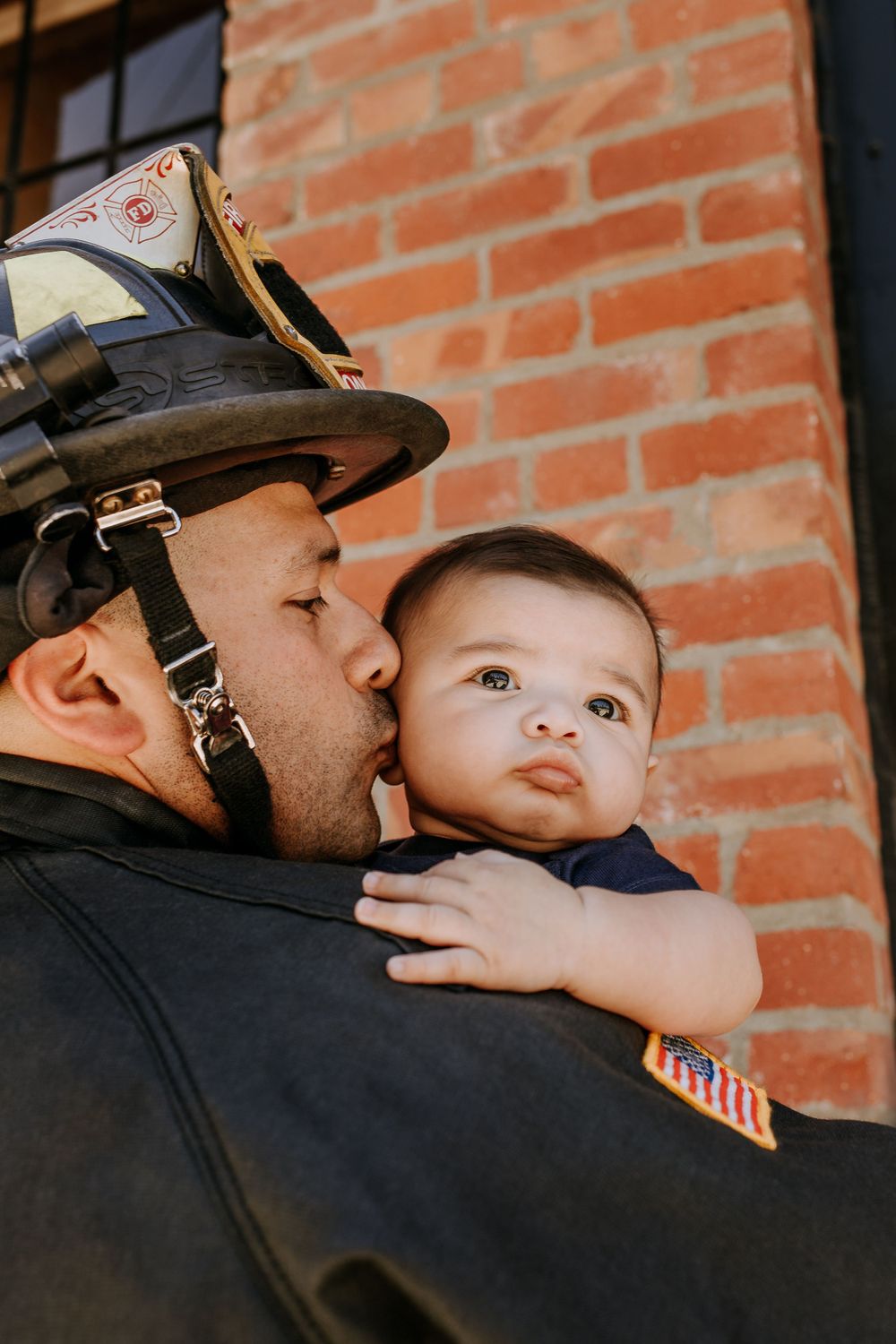 Firefighter in uniform helmet sharing tender moment with baby against brick wall.