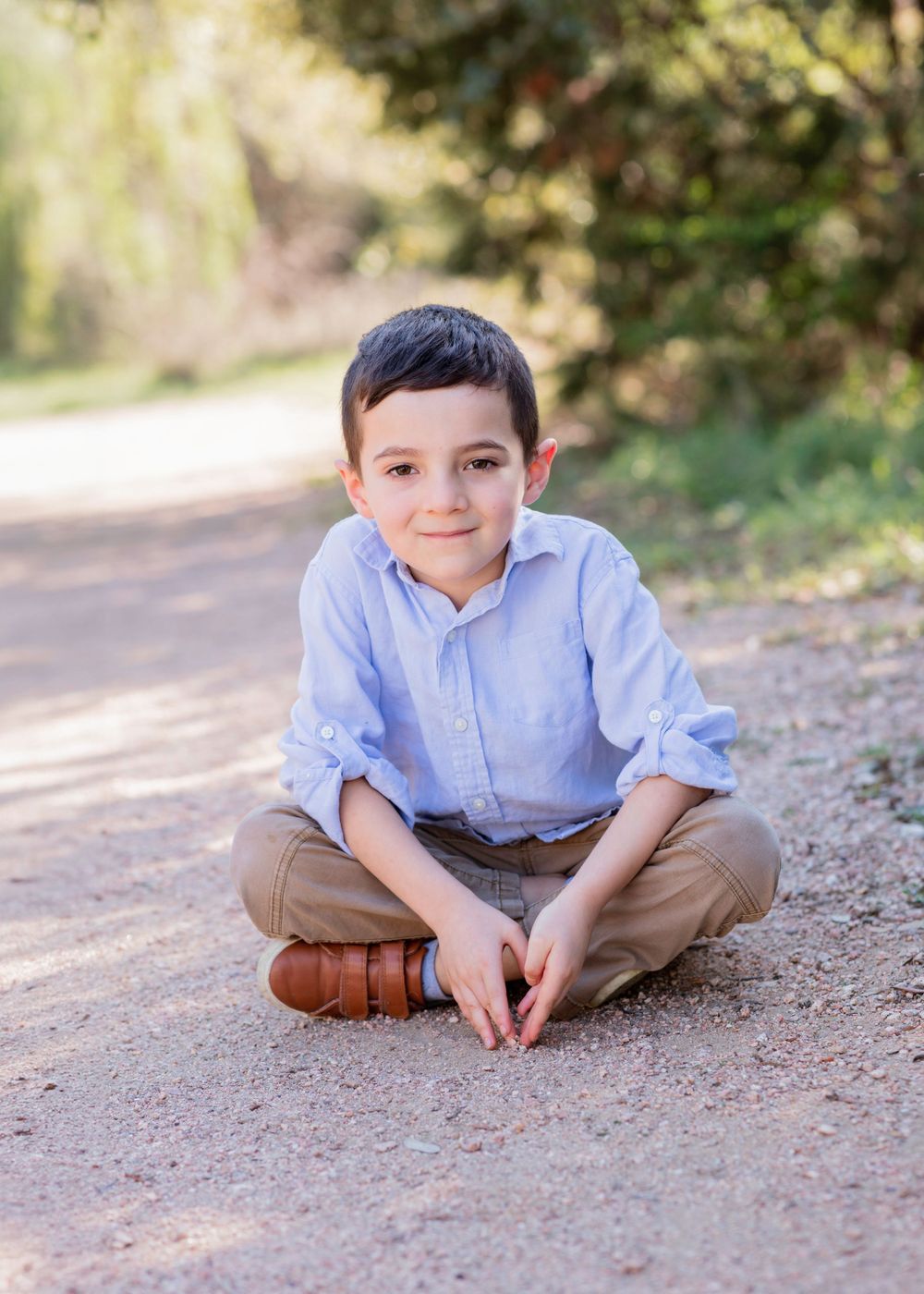 A child in a blue button-down shirt and khaki pants sits cross-legged on a dirt path surrounded by greenery.