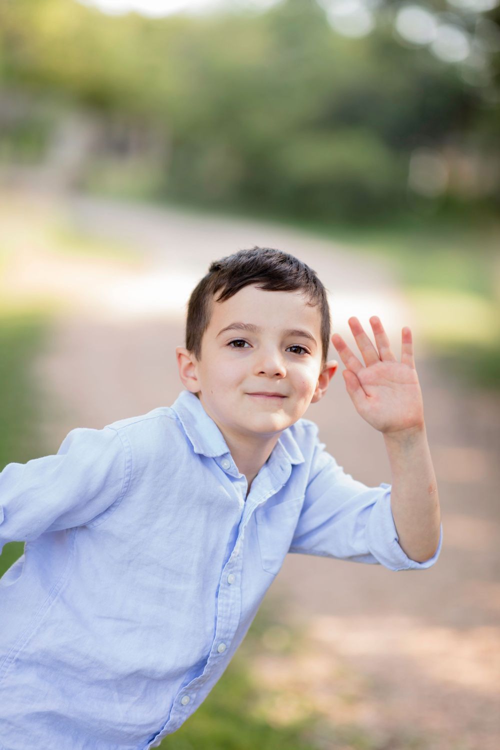 A young child wearing a light blue button-down shirt waves at the camera outdoors with a blurred natural background.
