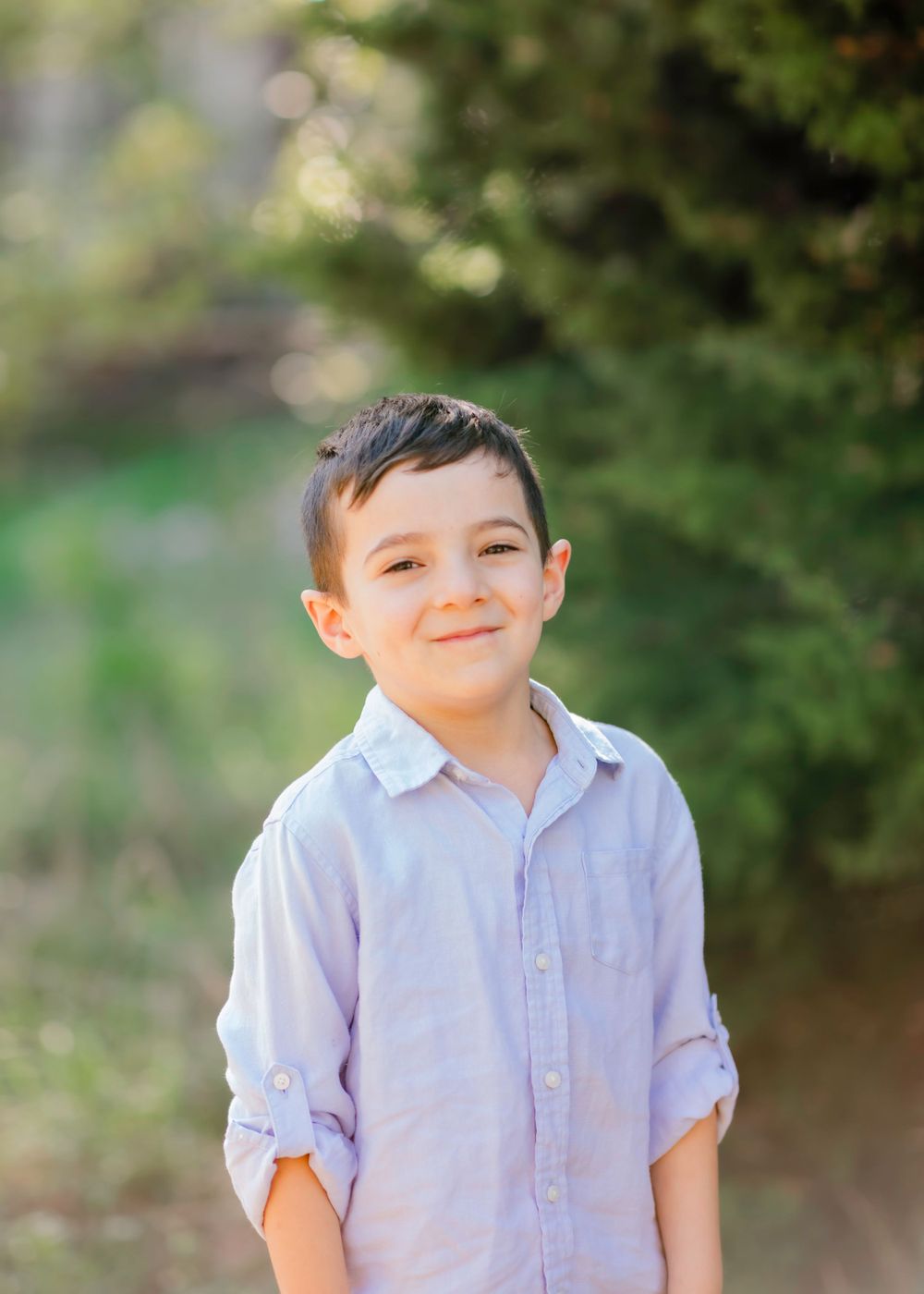 Young child wearing a light blue button-up shirt smiles outdoors with blurred green foliage in the background.