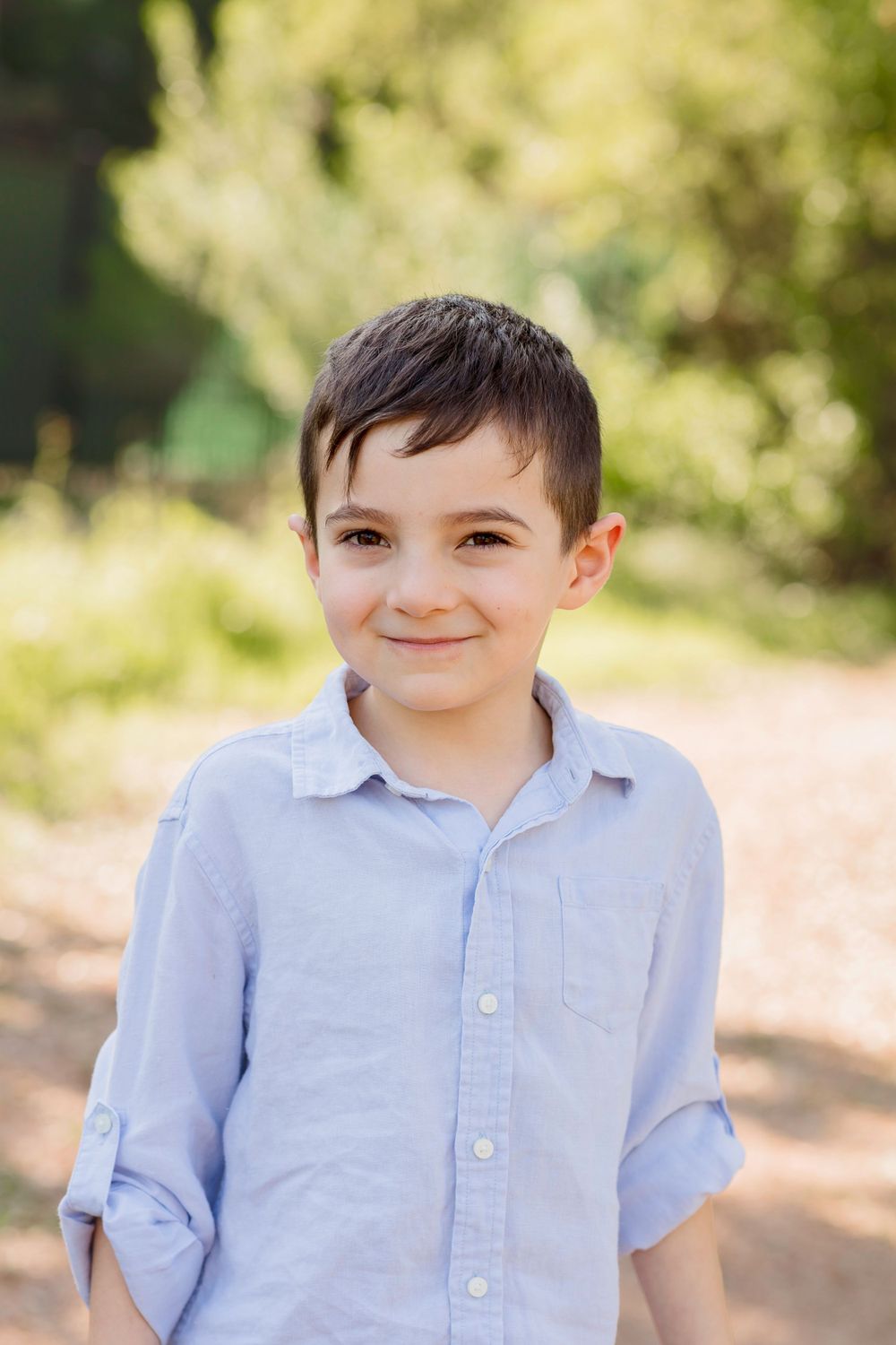 A young child in a light blue button-down shirt smiles warmly while standing outdoors on a sunny day.