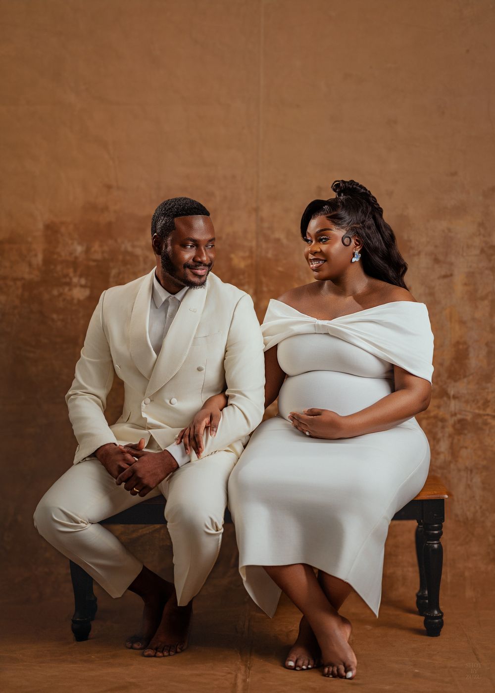 Elegant couple in matching cream outfits pose together against brown backdrop during maternity photoshoot.