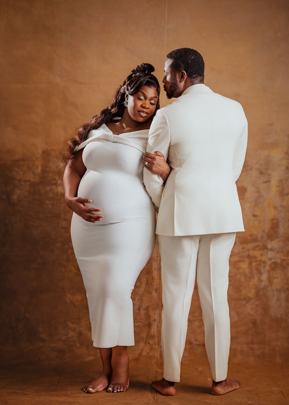 A tender maternity photo series showing a couple in white outfits against a rustic brown backdrop.