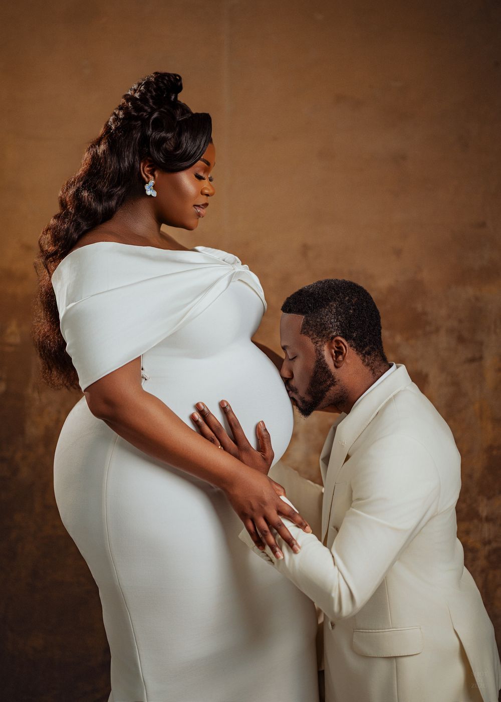 A couple in cream-colored outfits share an intimate moment during a maternity photoshoot against a brown backdrop.
