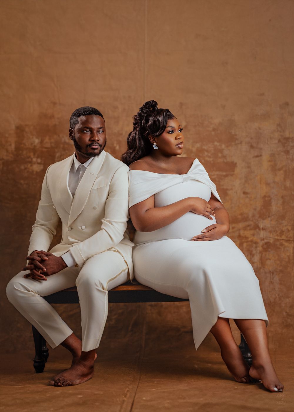 Couple in elegant white attire poses intimately on bench against textured brown wall in artistic photo series.