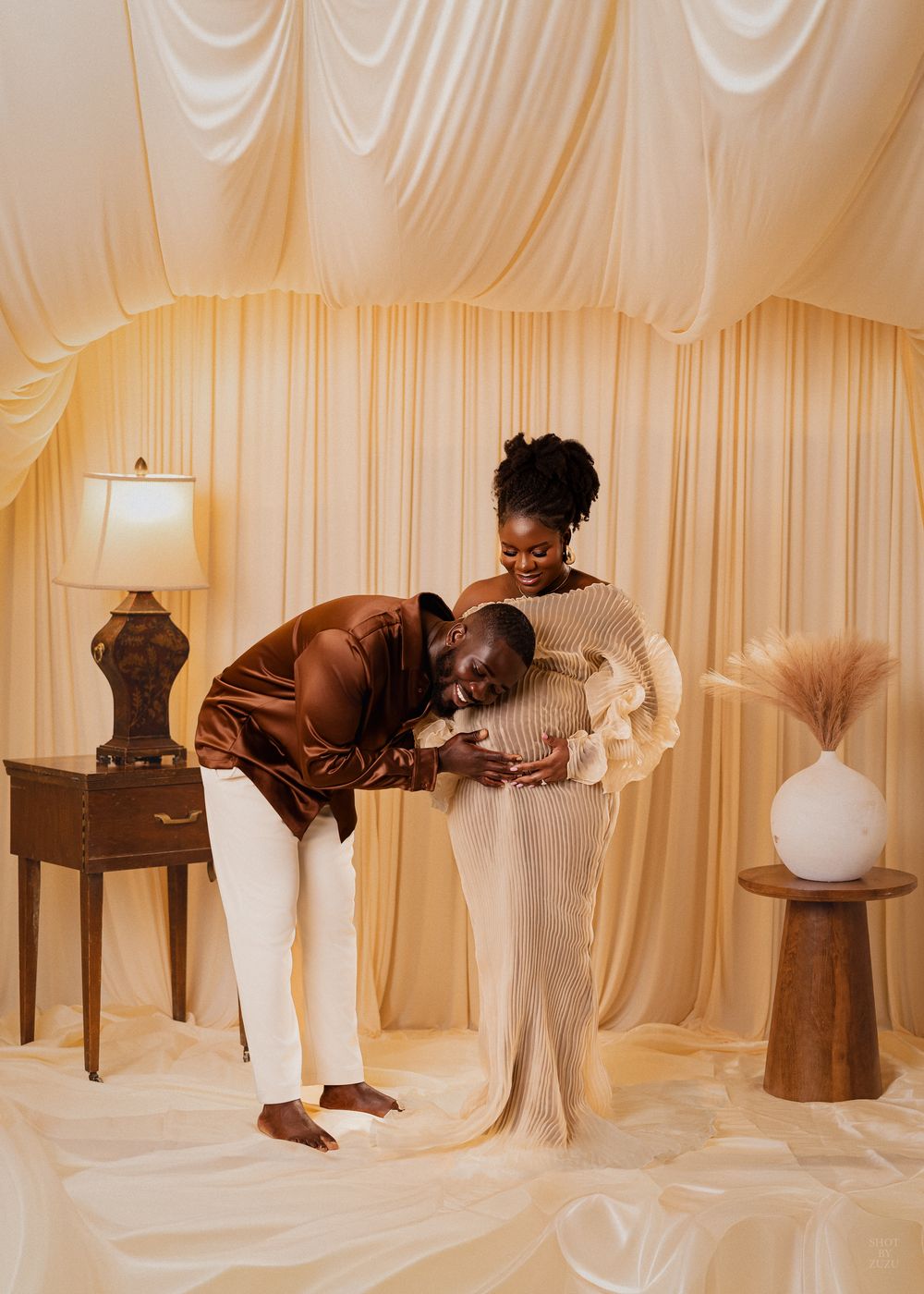 A tender moment captured in a warm-toned room with cream curtains, decorative lamp, and pampas grass decor.