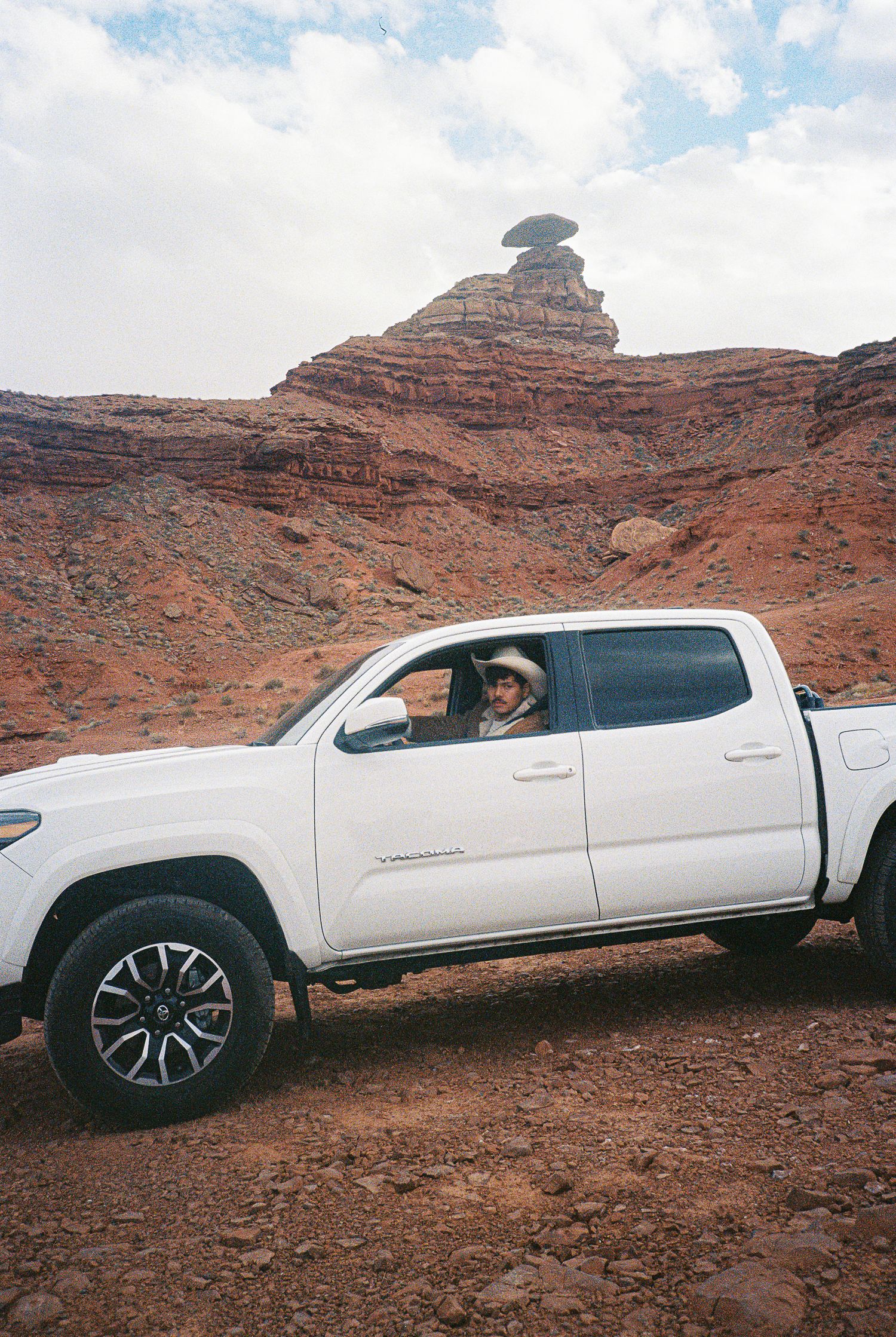 White pickup truck driving through red rock desert landscape with mountains in background.