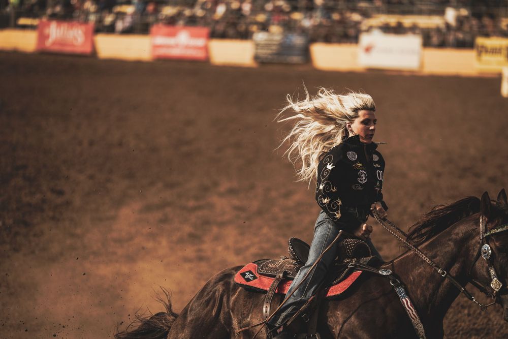 Rider with flowing hair galloping on horseback through dusty rodeo arena.