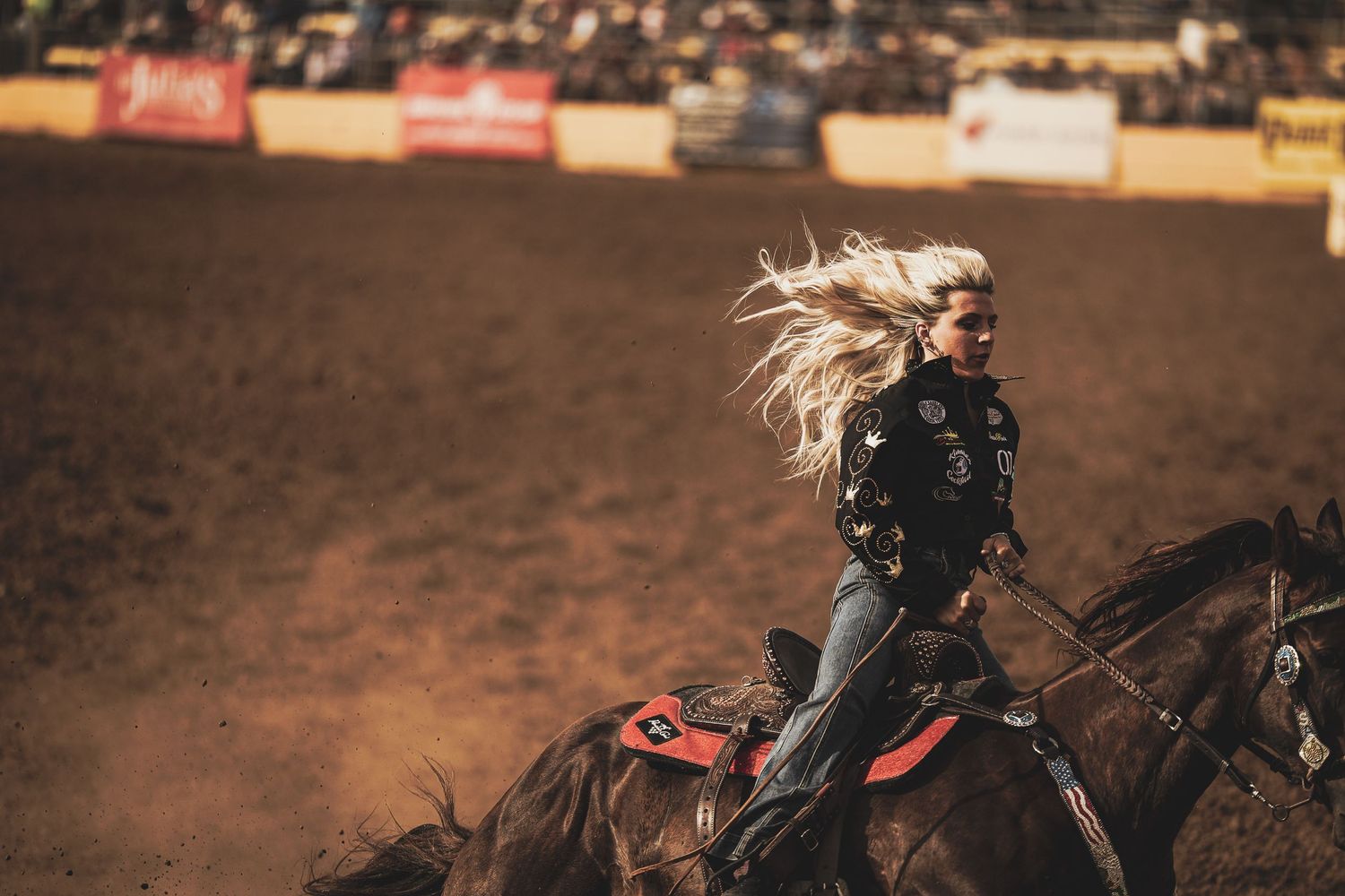 Rider with flowing hair galloping on horseback through dusty rodeo arena.