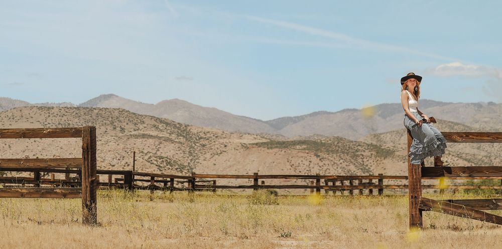 Woman in cowboy hat sitting on rustic wooden fence in vast desert landscape with mountains in background.