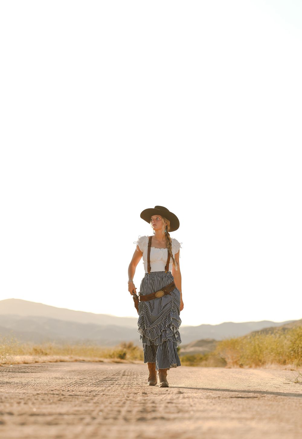 Woman in cowboy hat, white top, striped skirt, and boots walks down a desert road at golden hour.
